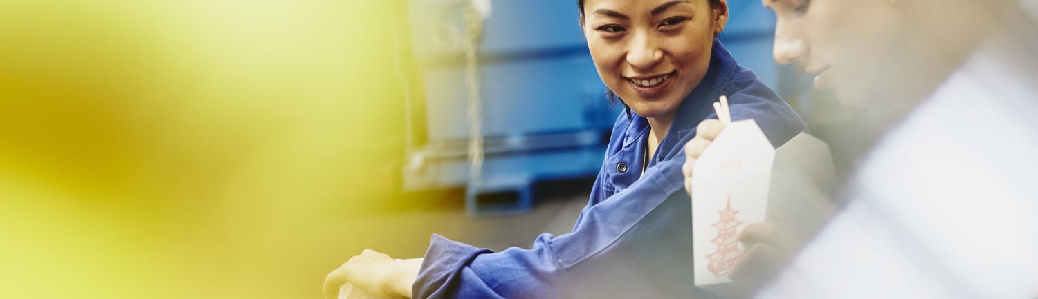 Male and female operator eating together. Food with chopsticks. Tech environment. Factory. Caucasian man and Asian woman. Primary color yellow. Secondary colors blue and white. 岗位外包